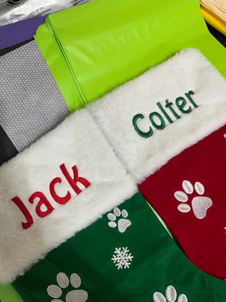 May include: Christmas stockings with white faux fur cuffs. One green stocking has the name "Jack" embroidered in red, and paw prints. Another green stocking has the name "Colter" embroidered in green. A third red stocking has paw prints.