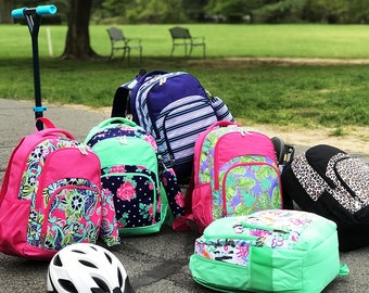 May include: A group of six colorful backpacks with various patterns, including floral, paisley, and tropical designs. The backpacks are arranged on a paved surface with a scooter in the background.