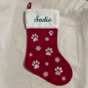 May include: Red Christmas stocking with white paw prints and snowflakes. The top of the stocking is white with the name "Sadie" embroidered in green. A red loop is attached for hanging.