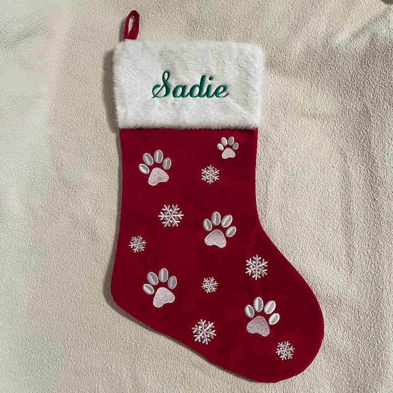 May include: Red Christmas stocking with white paw prints and snowflakes. The top of the stocking is white with the name "Sadie" embroidered in green. A red loop is attached for hanging.