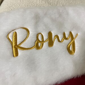 May include: A white Christmas stocking with a plush texture. The name "Rony" is embroidered in gold thread across the front. The stocking has a red backing and a cream-colored cuff.