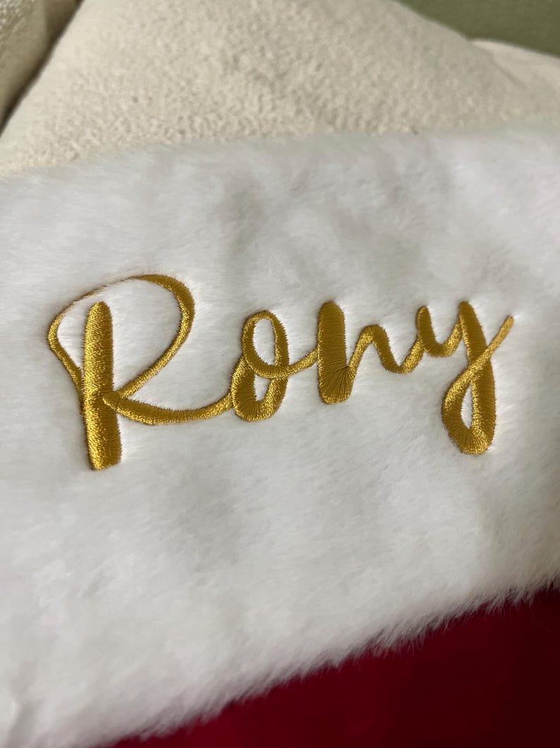 May include: A white Christmas stocking with a plush texture. The name "Rony" is embroidered in gold thread across the front. The stocking has a red backing and a cream-colored cuff.