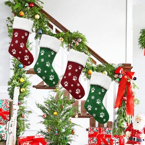 May include: Four Christmas stockings in red and green, adorned with white paw prints and snowflakes, hang on a staircase. Each stocking has a fluffy white cuff. The staircase is decorated with garland and ornaments.