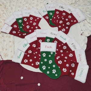 May include: A collection of Christmas stockings in red and green, adorned with white paw prints and snowflakes. Each stocking has a white cuff and a name embroidered in a contrasting color. The stockings are arranged on a textured surface.