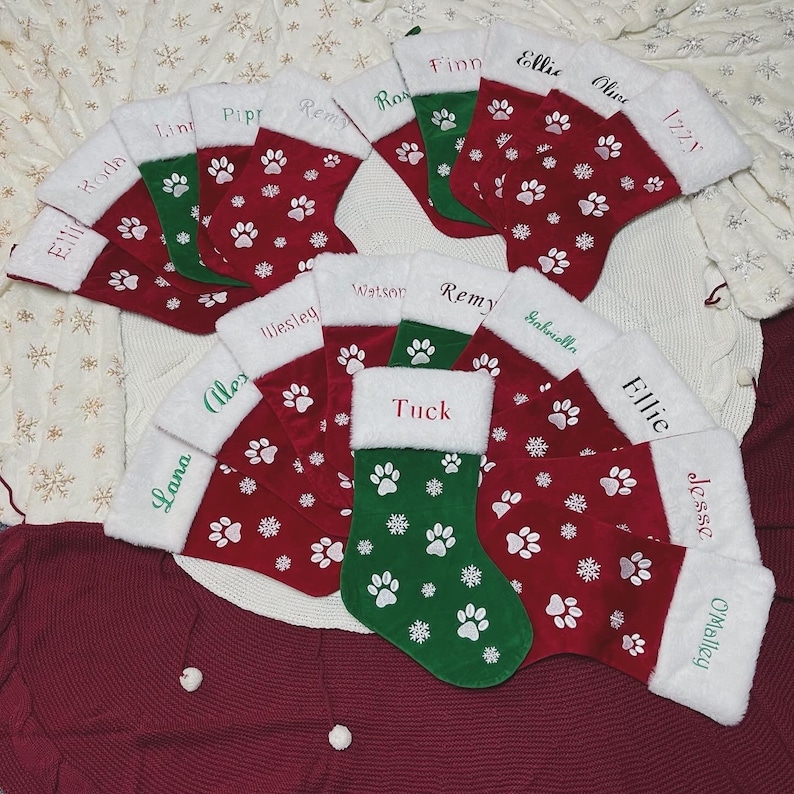 May include: A collection of Christmas stockings in red and green, adorned with white paw prints and snowflakes. Each stocking has a white cuff and a name embroidered in a contrasting color. The stockings are arranged on a textured surface.