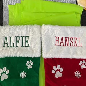 May include: Two Christmas stockings, one green and one red, with white faux fur cuffs. The green stocking has "ALFIE" embroidered in green, paw prints, and a snowflake. The red stocking has "HANSEL" embroidered in red, paw prints, and a snowflake.