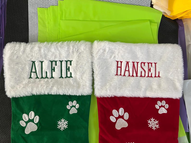 May include: Two Christmas stockings, one green and one red, with white faux fur cuffs. The green stocking has "ALFIE" embroidered in green, paw prints, and a snowflake. The red stocking has "HANSEL" embroidered in red, paw prints, and a snowflake.