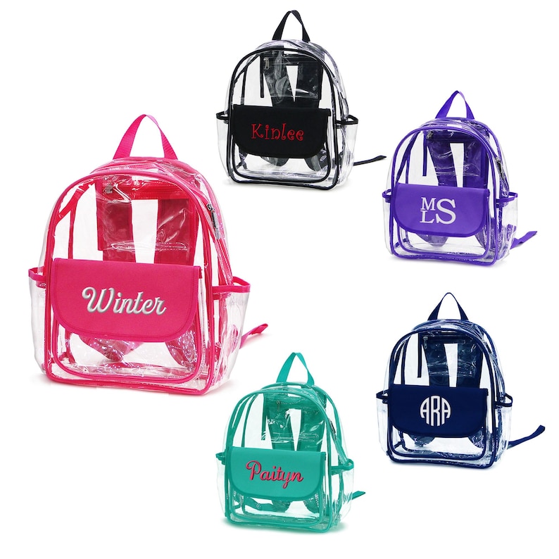 May include: Clear backpacks with colored accents and personalized text. The backpacks have a main compartment and a front pocket. The front pockets are in various colors, including pink, purple, teal, and navy. Each pocket has a name or initials embroidered on it.