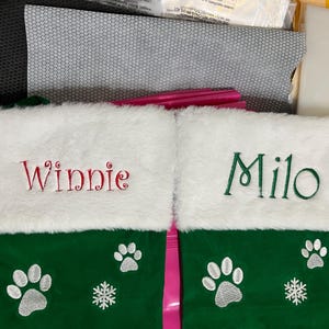 May include: Two green Christmas stockings with white faux fur cuffs. One stocking is embroidered with the name "Winnie" in red, the other with "Milo" in green. Each stocking features paw prints and a snowflake.