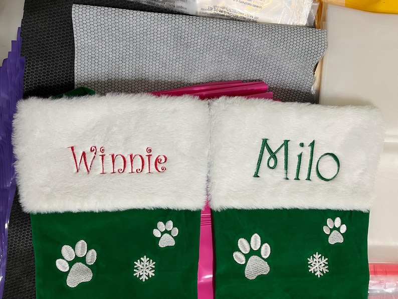May include: Two green Christmas stockings with white faux fur cuffs. One stocking is embroidered with the name "Winnie" in red, the other with "Milo" in green. Each stocking features paw prints and a snowflake.