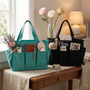 May include: Two tote bags, one teal and one black, sit on a wooden table. The teal bag has pockets filled with items like a perfume bottle and a notebook. The black bag has a photo and the name "Olivia" embroidered on it.
