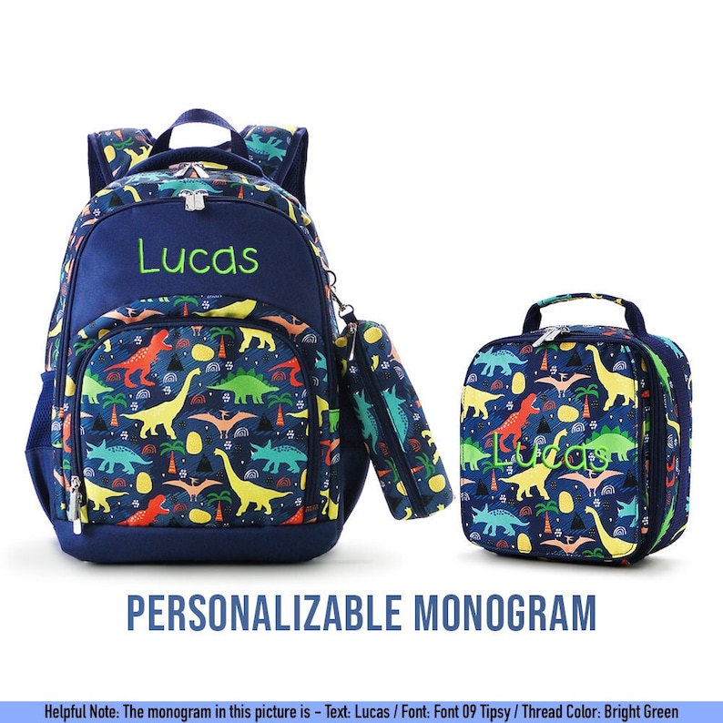 May include: A navy blue backpack, pencil case, and lunch bag set. The set features a colorful dinosaur pattern with the name "Lucas" embroidered in bright green. The text "PERSONALIZABLE MONOGRAM" is displayed below the set.