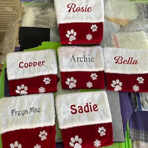 May include: Six Christmas stockings with white faux fur tops and red bottoms. Each stocking is personalized with a pet's name in cursive script. Paw prints and snowflakes decorate the red section, creating a festive holiday theme.