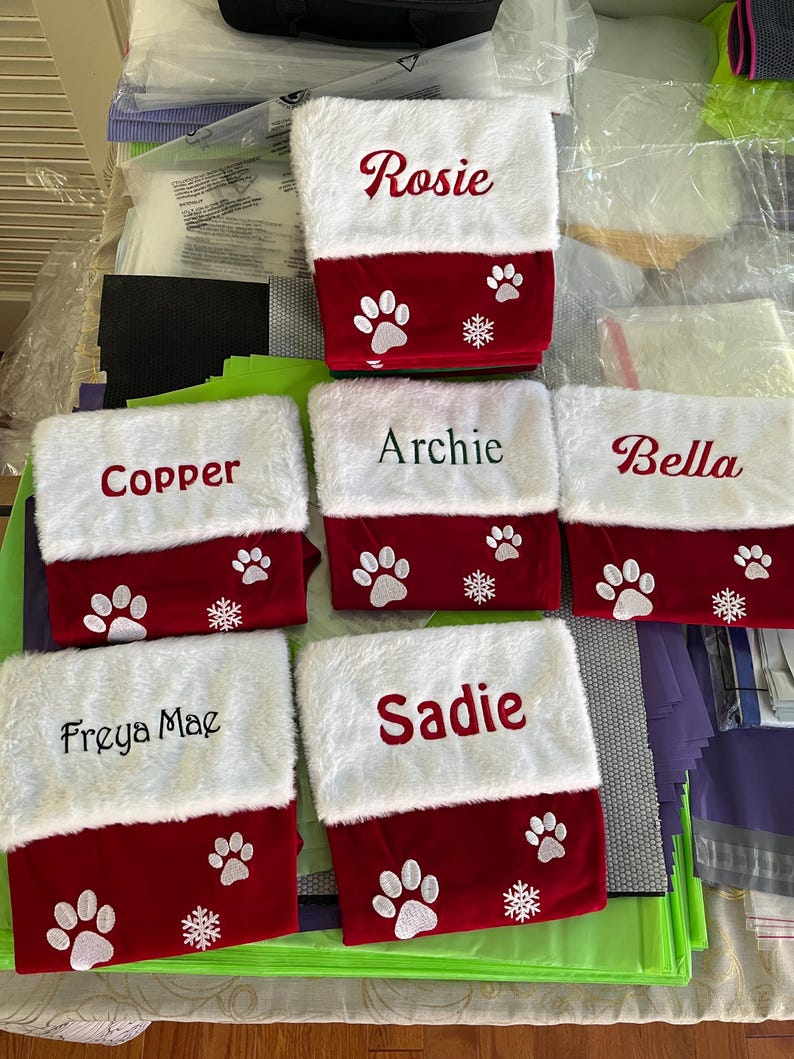 May include: Six Christmas stockings with white faux fur tops and red bottoms. Each stocking is personalized with a pet's name in cursive script. Paw prints and snowflakes decorate the red section, creating a festive holiday theme.