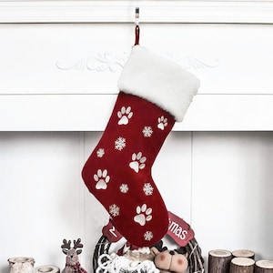 May include: A red Christmas stocking with white paw prints and snowflakes. The top of the stocking has a white faux fur trim. A red loop is attached for hanging. The stocking is hanging from a silver hook.