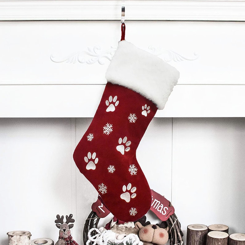 May include: A red Christmas stocking with white paw prints and snowflakes. The top of the stocking has a white faux fur trim. A red loop is attached for hanging. The stocking is hanging from a silver hook.