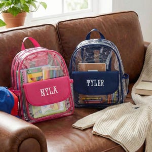 May include: Two clear backpacks with colorful contents sit on a brown leather couch. One backpack is pink with the name "NYLA" embroidered on the front pocket. The other is blue with "TYLER" embroidered on the front pocket.