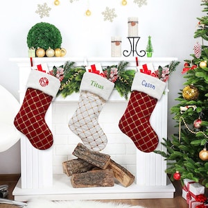 May include: Three Christmas stockings with personalized embroidery. The stockings are red and white with a diamond pattern. The names on the stockings are "ASM", "Titus", and "Cason".