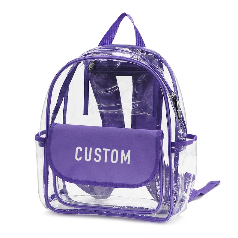 May include: Clear backpack with purple trim and accents. Features a front flap with the word "CUSTOM" in white. Includes a top handle, adjustable straps, and a zippered main compartment. Perfect for school or events.