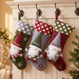 May include: Three Christmas stockings hanging on hooks, each featuring a gnome applique with a white beard and a pointed hat. The stockings are green, gray, and red, decorated with pom-poms, stars, and embroidered names Laura, Kathy, and Karter.