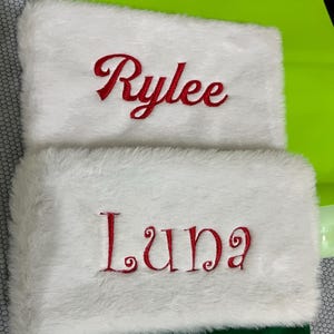 May include: Two white Christmas stockings with red embroidered names. The top of each stocking has a fluffy white trim. One stocking says "Rylee" and the other says "Luna". The bottom of the stockings are green.