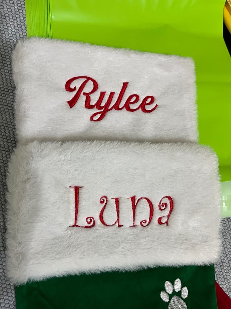 May include: Two white Christmas stockings with red embroidered names. The top of each stocking has a fluffy white trim. One stocking says "Rylee" and the other says "Luna". The bottom of the stockings are green.