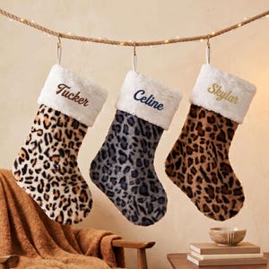 May include: Three plush Christmas stockings hanging from a rope. The left stocking has a brown leopard print and the name Tucker. The middle stocking has a gray leopard print and the name Celine. The right stocking has a brown leopard print and the name Skylar.