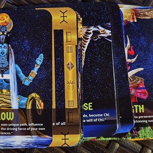 May include: A deck of oracle cards with a blue-skinned deity on the first card, labeled "FLOW". The cards have gold accents and feature various illustrations and text. The cards are stacked on a dark surface.