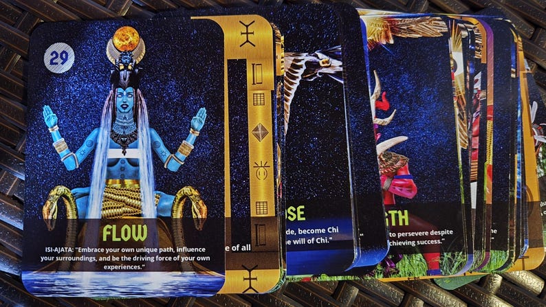 May include: A deck of oracle cards with a blue-skinned deity on the first card, labeled "FLOW". The cards have gold accents and feature various illustrations and text. The cards are stacked on a dark surface.