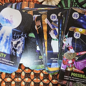 May include: A deck of oracle cards with colorful illustrations and text. The cards feature images of a waterfall, a figure with a bow and arrow, and other symbolic designs. The word "Purity" and "Possibility" are visible on the cards.
