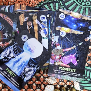 May include: A collection of oracle cards with celestial and mythological imagery. The cards feature illustrations of figures, celestial bodies, and text such as "Fortune," "Purity," and "Possibility." The cards are on a patterned fabric.