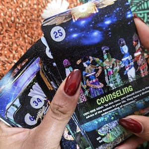 May include: A deck of oracle cards with a black background and images of figures in colorful clothing. The card in focus has the word "COUNSELING" and the number "25". The cards are held in a person's hand.