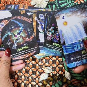 May include: A deck of oracle cards with colorful illustrations and text. The cards feature images of waterfalls, figures, and celestial bodies. The word "FORTUNE" is visible on one card. The deck is displayed on a patterned fabric.