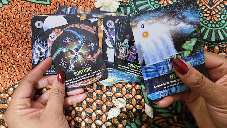 May include: A deck of oracle cards with colorful illustrations and text. The cards feature images of waterfalls, figures, and celestial bodies. The word "FORTUNE" is visible on one card. The deck is displayed on a patterned fabric.
