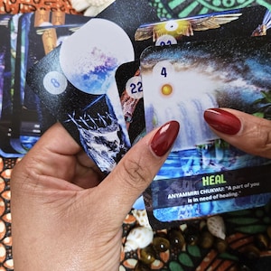 May include: A deck of oracle cards featuring celestial and nature-themed imagery. The cards are held in a person's hands, with the word "HEAL" visible on one card. The deck includes cards with numbers and illustrations of waterfalls and moons.