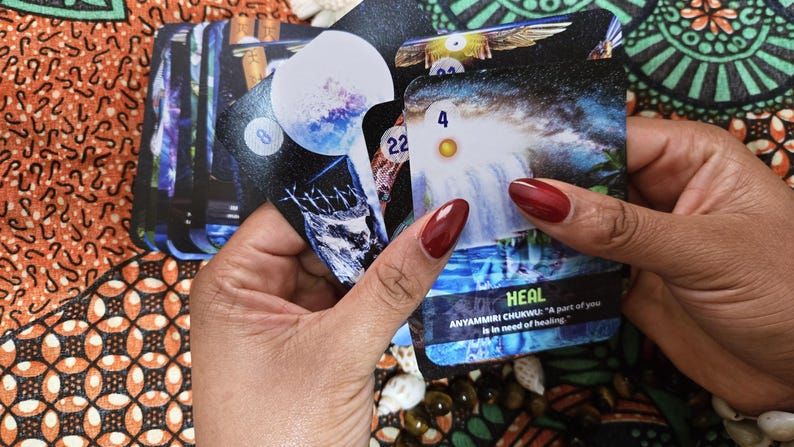 May include: A deck of oracle cards featuring celestial and nature-themed imagery. The cards are held in a person's hands, with the word "HEAL" visible on one card. The deck includes cards with numbers and illustrations of waterfalls and moons.