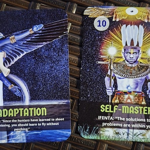 May include: Two oracle cards with illustrations and text. One card features a blue bird-like figure with the word "ADAPTATION" and a quote. The other card shows a figure holding staffs, with the word "SELF-MASTERY" and a quote.