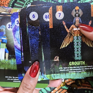 May include: A deck of tarot cards featuring colorful illustrations and numbers. One card displays the word "GROWTH" with an image of a woman with wings. Other cards show figures and celestial designs. The cards are held in a person's hand.