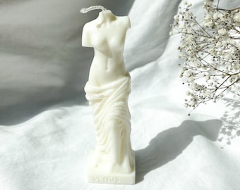 Könnte beinhalten: Elfenbeinfarbene Kerze in Form der Venus von Milo Statue. Die Kerze zeigt den Oberkörper einer Frau, in Stoff gehüllt, mit dem Wort "VENUS" an der Basis. Ein weißer Docht ragt heraus. Die Kerze steht auf einer weißen Oberfläche.