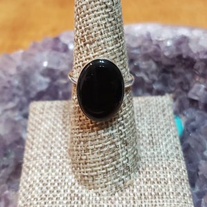 Peut inclure: Une bague en argent avec une grosse pierre noire ovale polie. La bague est présentée sur un présentoir à bagues beige texturé. L'arrière-plan comprend une surface en bois floue et un cristal violet.