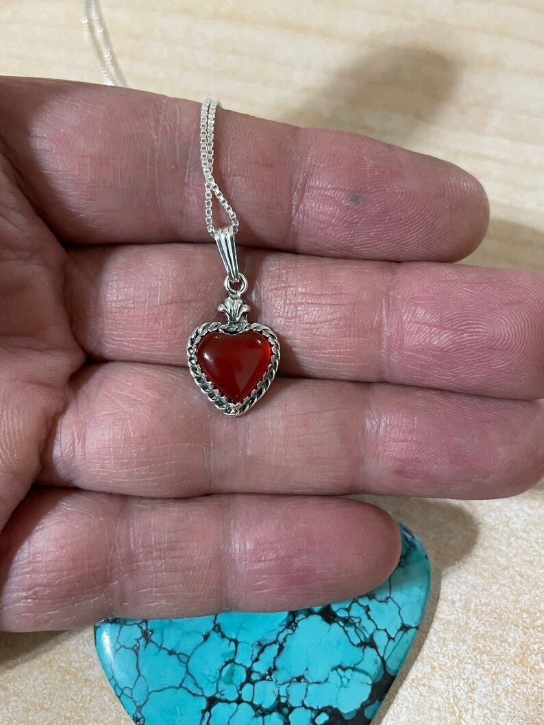 May include: A silver heart-shaped pendant necklace with a red carnelian stone. The necklace is on a silver chain.