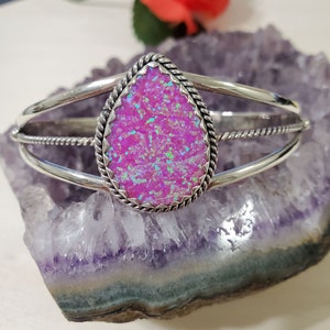 May include: A silver bangle bracelet with a pink opal teardrop-shaped stone set in a silver bezel. The stone has a sparkly, iridescent finish.
