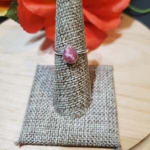 Puede incluir: Un anillo de plata con una piedra preciosa rosa y blanca. El anillo se muestra en un soporte de anillo de tela marr&oacute;n.