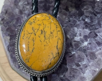 May include: A bolo tie featuring a yellow and black patterned stone set in a silver oval frame. The bolo tie is on a black braided cord.