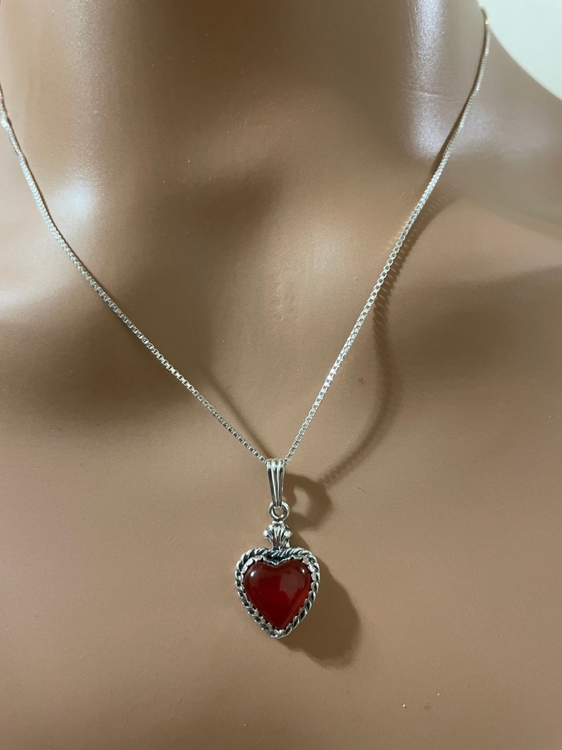 May include: A silver chain necklace with a red heart pendant. The heart is set in a silver frame with a detailed design.