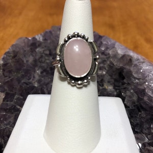 May include: A silver ring with a light pink oval-shaped gemstone set in a decorative silver bezel. The ring is on a white ring display stand.