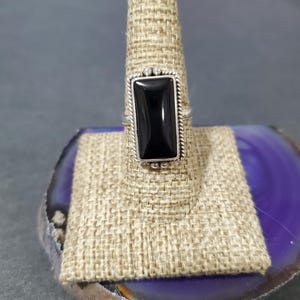 May include: A silver ring with a black rectangular gemstone set in a bezel setting. The ring is on a display stand.