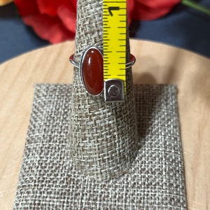 May include: A silver ring with a red carnelian oval gemstone. The ring is displayed on a fabric ring holder with a measuring tape in the background.