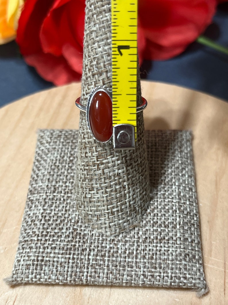 May include: A silver ring with a red carnelian oval gemstone. The ring is displayed on a fabric ring holder with a measuring tape in the background.