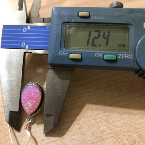May include: A pink opal gemstone earring with a silver setting, measured at 12.4 mm in diameter using a digital caliper.